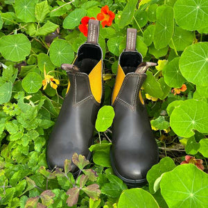 Blundstone Brown Chelsea boots with yellow accents and tags on a bed of green leaves and flowers 